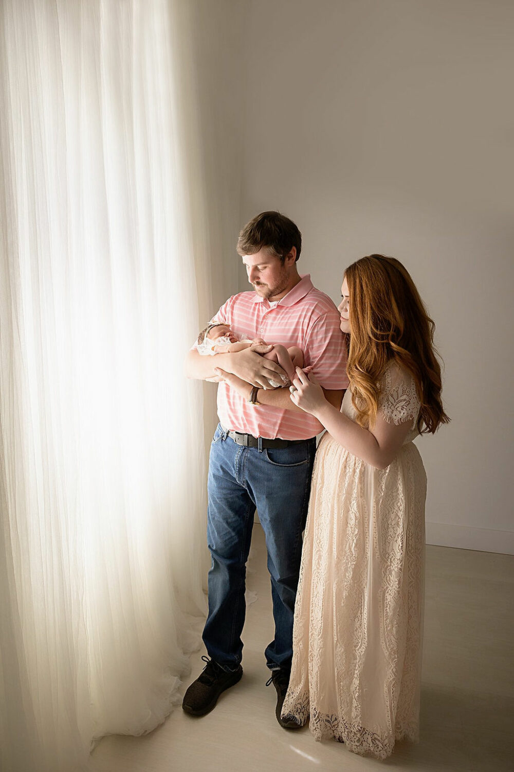 Man holding his newborn daughter in his arms as his wife stands behind him, and they are both looking at their baby for their neutral in studio newborn session, taken in Westhampton, New Jersey.