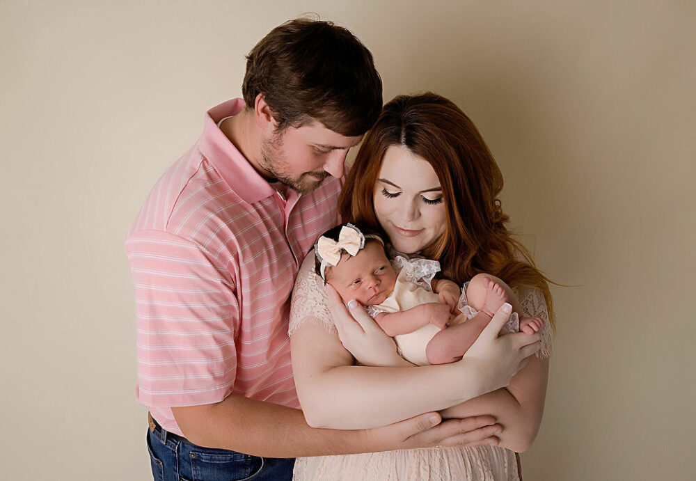 Man embracing his wife as she holds their newborn daughter in her arms as they both look at her smiling for their newborn family photos taken during their Neutral in-studio newborn session in Southampton, New Jersey.