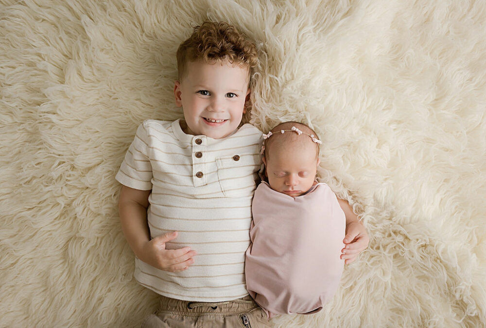Aerial picture of a toddler boy, smiling at camera as he hugs his newborn sister as they both lay on newborn photography blankets, taking for her pink in-studio newborn session in freehold, New Jersey.