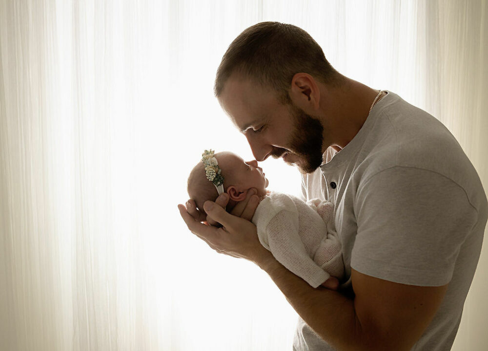 A father holding his newborn daughter in his hands, touching noses with her for her pink in-studio newborn session, taken in Mount Holly, New Jersey.