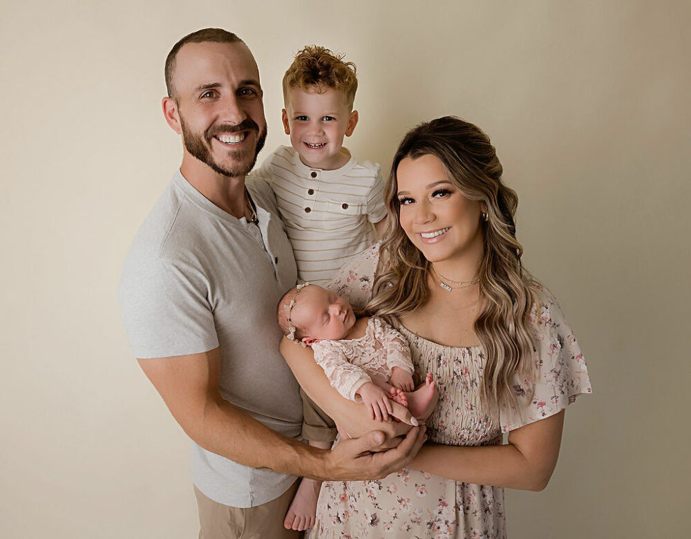 Man and woman smiling at camera while holding their infant daughter and toddler son in their arms for baby pics taken during their daughter’s newborn session taken in Eastampton, New Jersey.