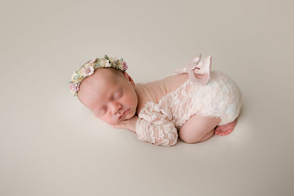 Infant girl, sleeping on tummy wearing newborn outfit with bow and flowery headband for her newborn photography, taken in studio for her pink in-studio newborn session in Southampton, New Jersey.