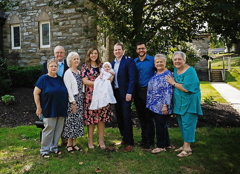 Family portrait of new parents and their extended family posing and smiling at camera as a stand in front of a church while there newborn girl in baptism outfit is in mom's arms for their baby baptism a family session in Hamilton, New Jersey.