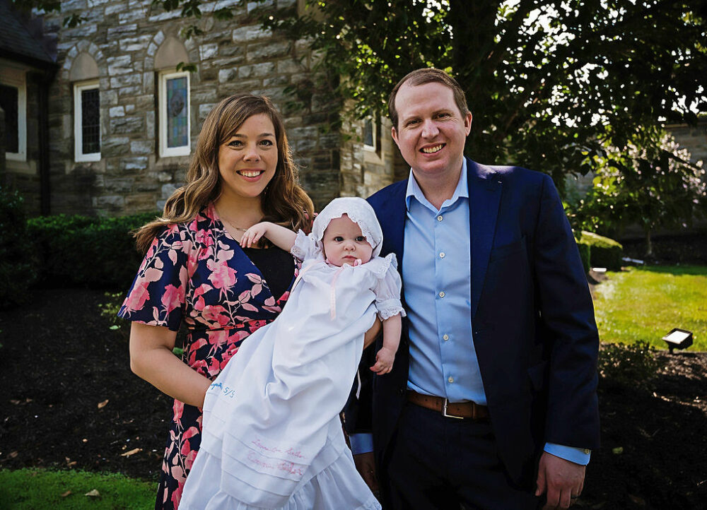 Man and woman smiling at camera as women hold her newborn daughter as they wear their Sundays best for her baby baptism a family session in Crosswicks, New Jersey.