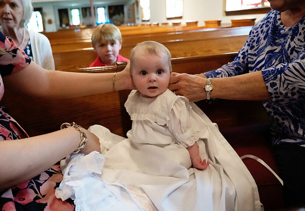 Newborn girl wearing baptism clothing looking at camera sitting on church pews along with her mother and extended family for her baby baptism and family session taken in Westampton, New Jersey.