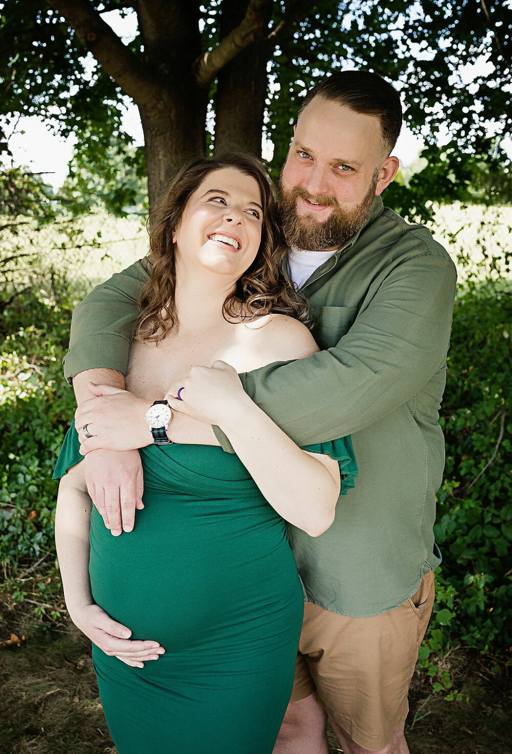 Man, standing behind, woman, holding her as she looks at him and hold her belly standing outdoors for their fall maternity photography near me in Westampton, New Jersey.