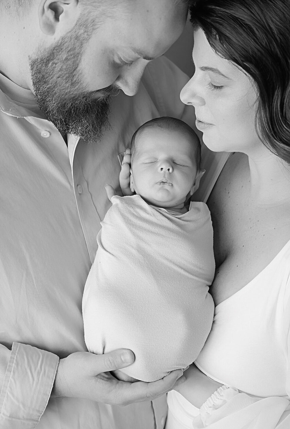 Black-and-white close-up of a man and woman, touching foreheads, and holding their newborn son in between them as he sleeps, and is swaddled for their newborn picture ideas taken in Cinnaminson, newJersey.