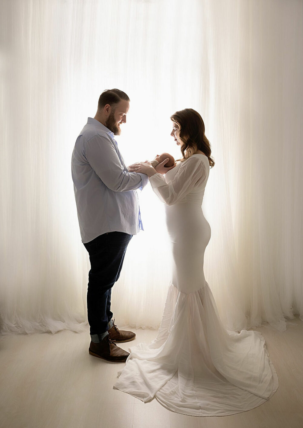 Man and woman facing each other, while they both hold their newborn sun in between them, facing each other, standing against a backlit background for their fall and studio maternity session in Bordentown, New Jersey.
