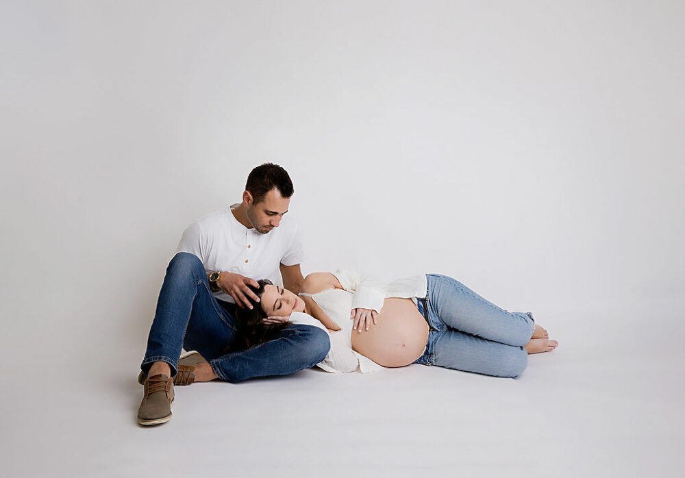 Man sitting on floor with a woman laying down and resting her head on his lap as she holds her tummy, he's loving looking at her, for a white and bright maternity session in Southampton, New Jersey.