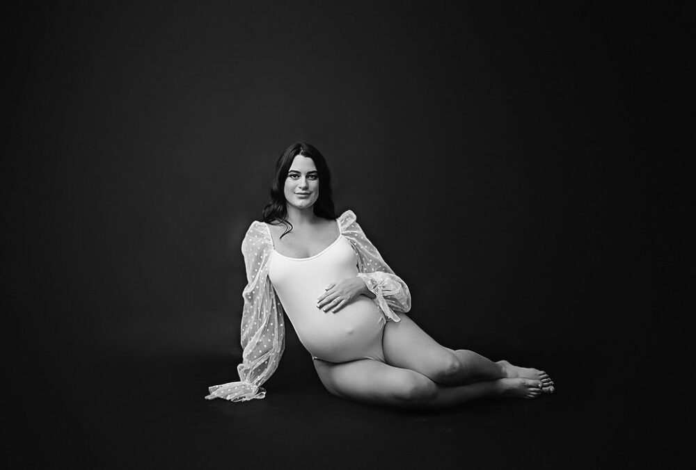 Woman sitting on the floor leaned on one side using her right hand as support while her left and is on her belly wearing ballet maternity outfit looking at camera for her white and bright maternity session in Westampton, New Jersey.