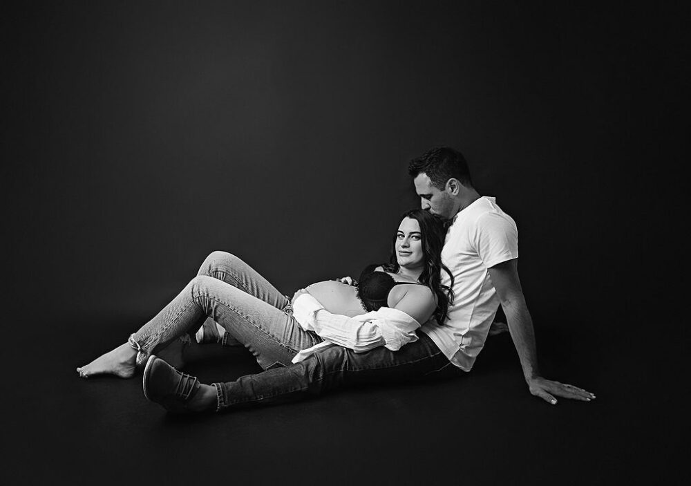 Couple maternity portrait of a man sitting on floor as his wife is laying between his legs as he's kissing her head taken for their in-studio maternity session in Eastampton, New Jersey.