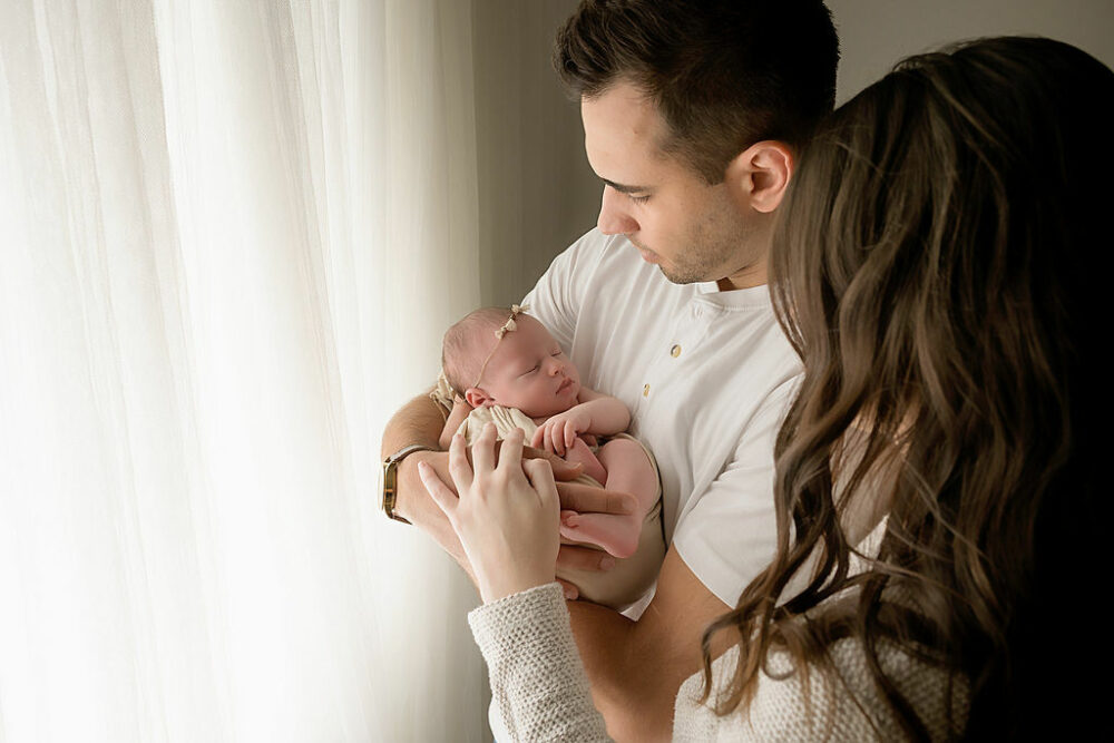 Man and woman looking down at their newborn against a light and bright background for there in studio White and bright newborn session in Burlington, New Jersey.