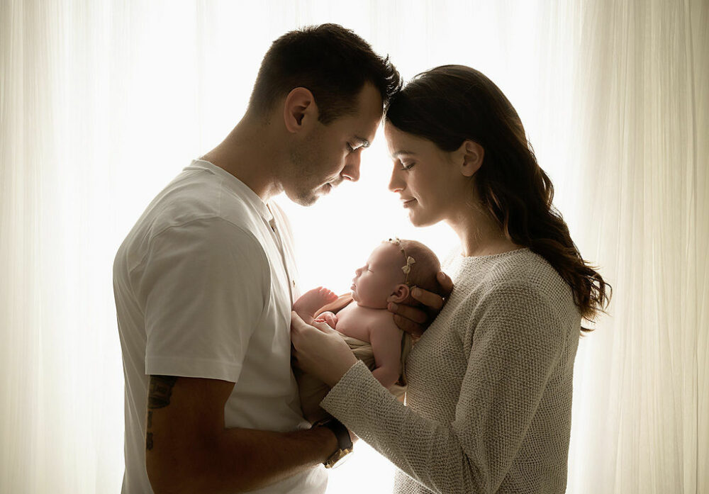 Man and woman facing each other and touching foreheads as they hold their newborn daughter in between them for their in studio infant photo shoot taken in Hamilton, New Jersey.