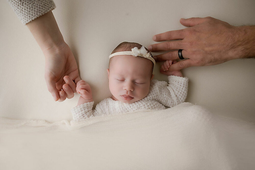 Infant portrait of baby girl sleeping on her back wearing a knit outfit and matching headband holding her parents hands as she sleeps soundly taken during her in studio white and bright newborn session in Mount Holly, New Jersey.