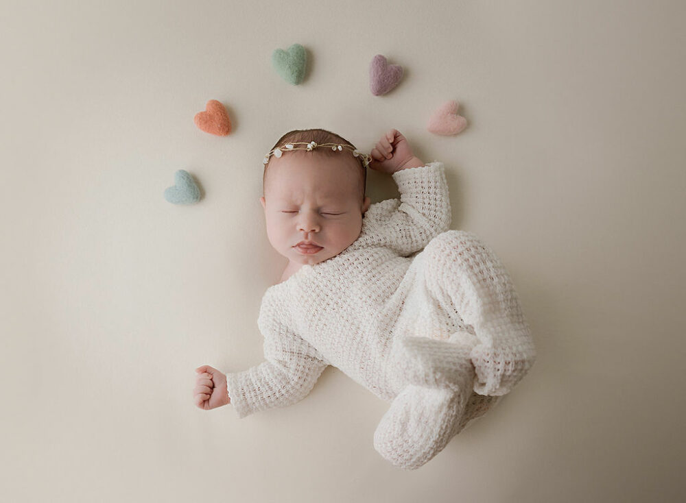 Newborn photography of infant girl laying on the back with hands on her side wearing that outfit adorned with colorful felt hearts for her white and bright newborn session taken in Cherry Hill, New Jersey.