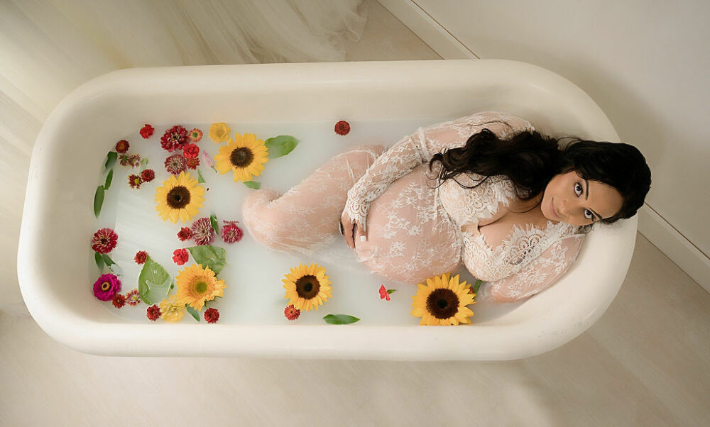 Woman laying in bathtub and looking up at camera while holding her belly, wearing maternity dress for her in studio milk, bath maternity session taken in Westhampton, New Jersey.