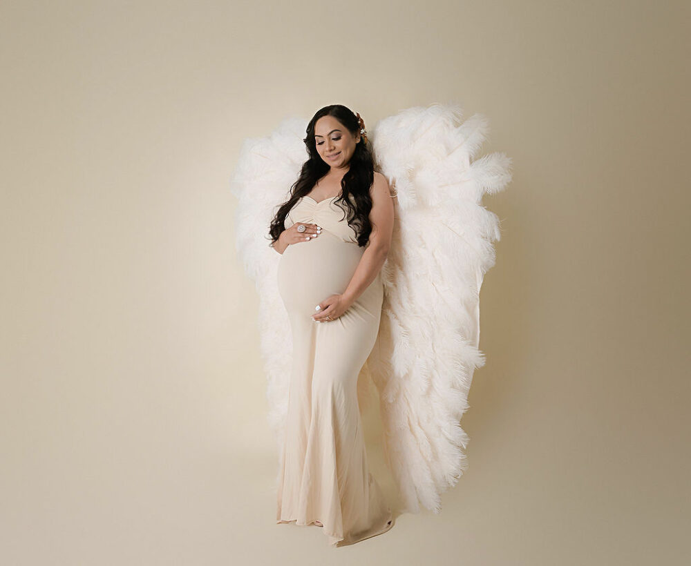 Woman holding her belly and looking down wearing maternity dress and angel wings for her in studio milk bath maternity session take in Cherry Hill, New Jersey.