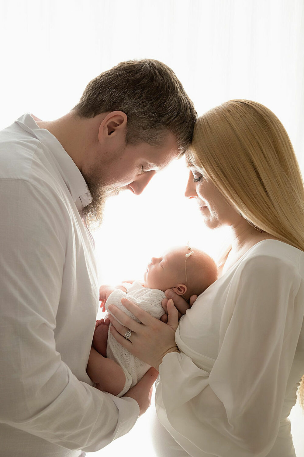 Close-up of man and woman looking down at their baby as they hold her between them, touching foreheads for their in studio tan maternity session