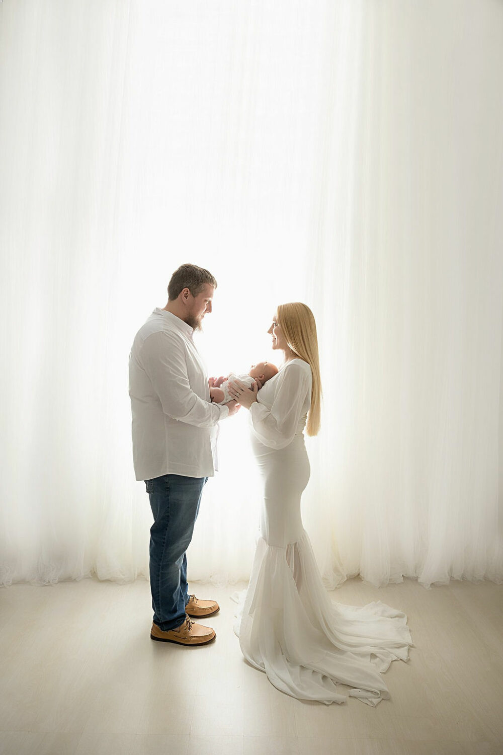 Man and woman facing each other while they both hold their newborn between them and looking at each other standing against a bright background for their classic newborn session taken in Westampton, New Jersey.