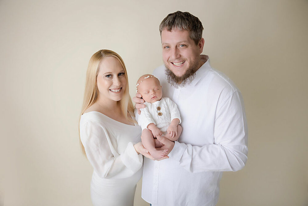 Man and woman standing next to each other looking and smiling a camera while holding their newborn together for their baby portraits taken in Eastampton, New Jersey.