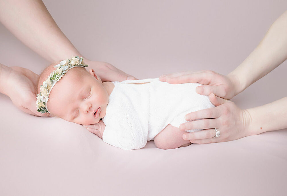 Infant girl laying on her tummy wearing outfit matching headband sleeping as her parents hold her sweetly for her classic newborn session taken in Princeton, New Jersey.