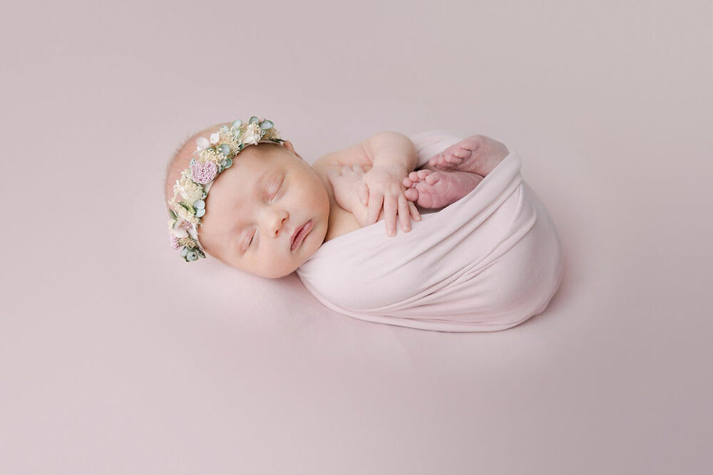 Infant girl partially wrapped and laying on her back sleeping and wearing headband for her newborn portrait taken and professional photography studio for her classic newborn session in Somerville, New Jersey.