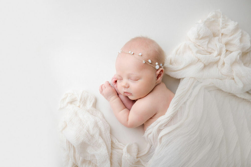Infant sleeping on side covered with blanket wearing headband for her classic newborn session taken in Bridgewater, New Jersey.