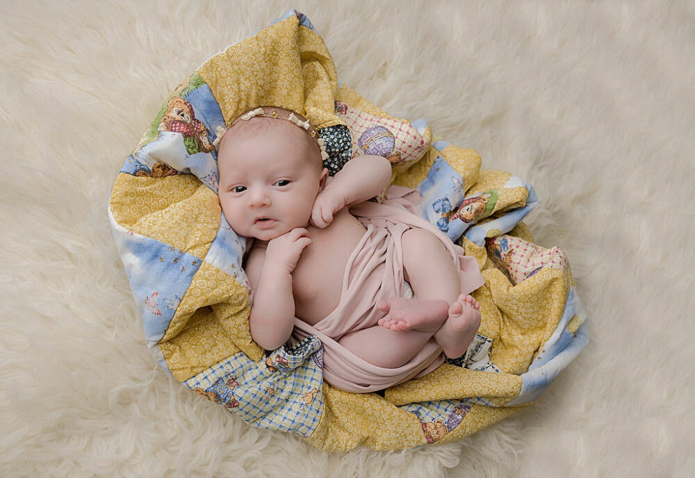 Infant girl awake and laying on back on family heirloom against a picture blanket wearing a headband for her newborn photography session in Burlington, NJ.