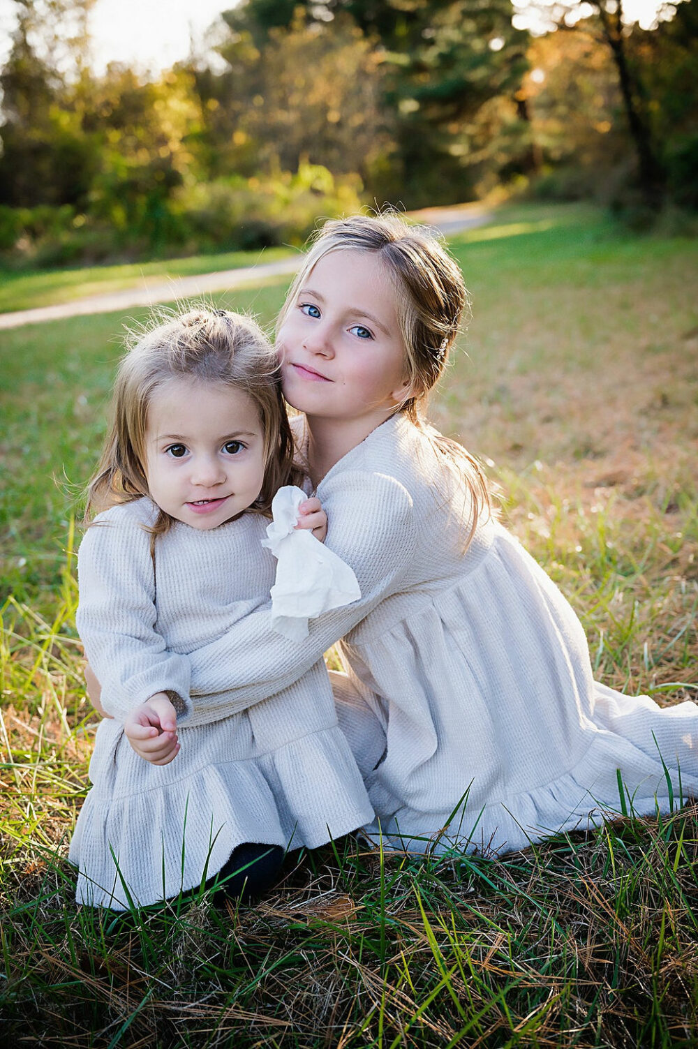 Two toddler girls wearing matching dresses sitting on grass and posing for the camera for their Berlin Park family session taken in South Jersey.