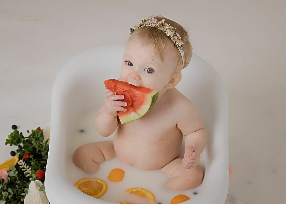 One year old girl biting watermelon slice as she sits in tiny tub for her fruit medley first birthday themes in Westampton, New Jersey.