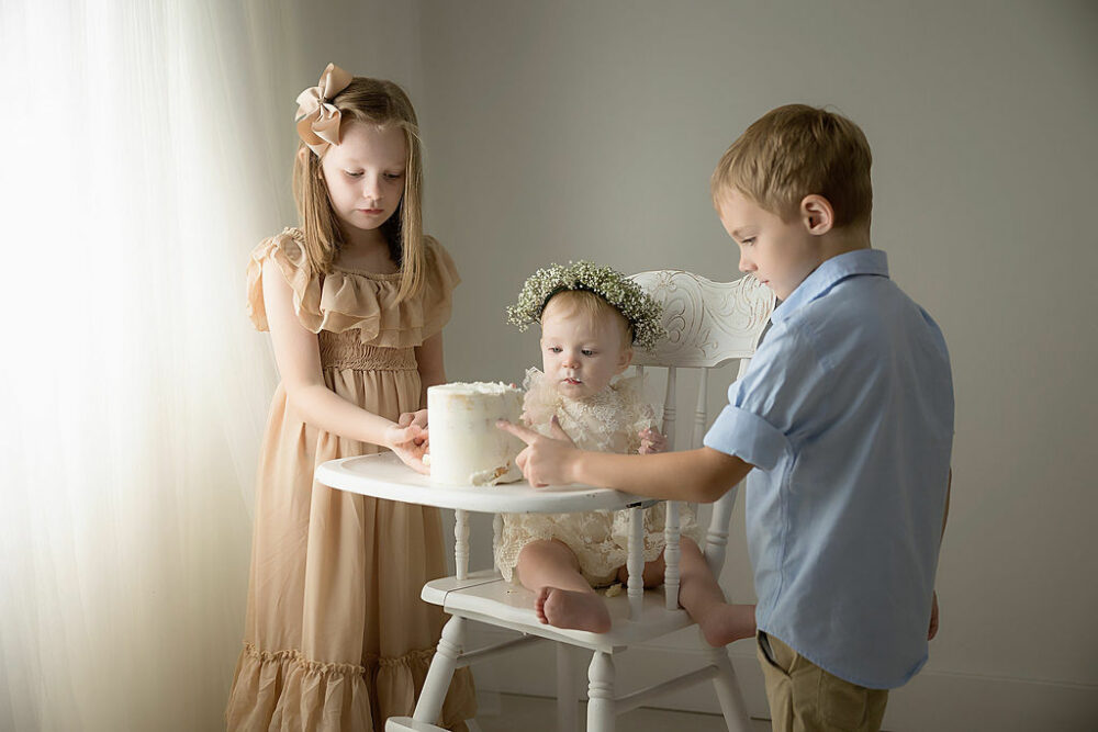 Sibling portrait of big brother and big sister eating first birthday cake with their one year old sister for her cake smash session taken in Easthampton, New jersey.
