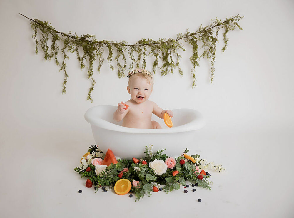 Toddler girl smiling and looking at camera as she sits in bathtub photography prop holding fruit well photo Saint Isidore with greenery and flowers for her fruit medley first birthday session in Cherry Hill, NJ.