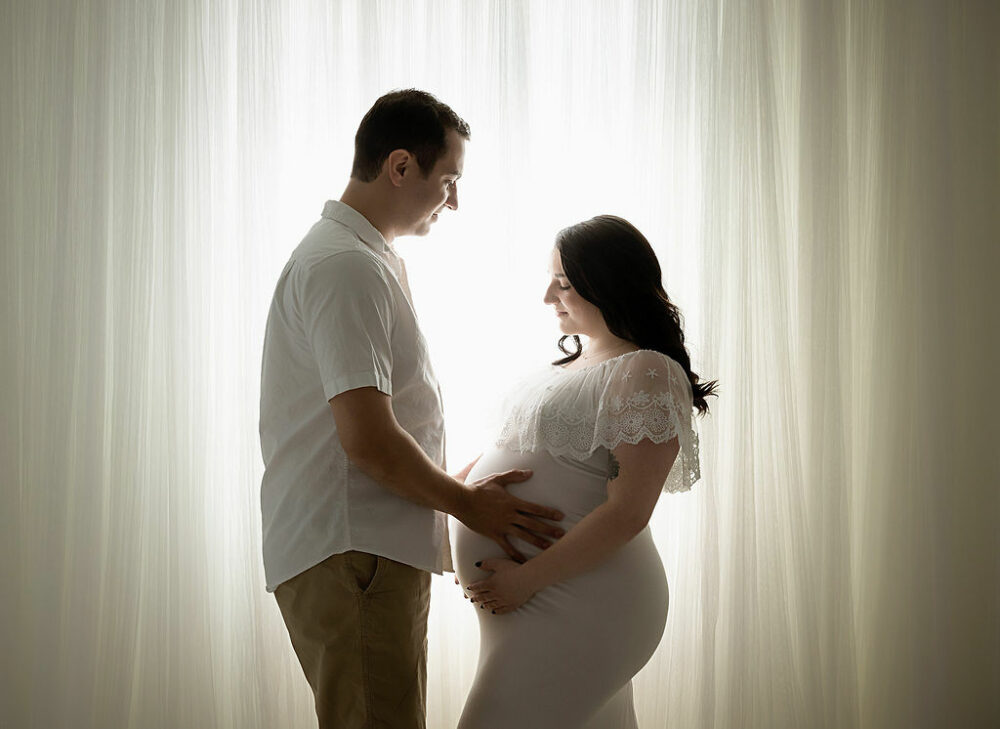 man and woman facing each other while holding her belly against a light and bright background for their couple maternity photos taken in studio in Burlington, New Jersey.