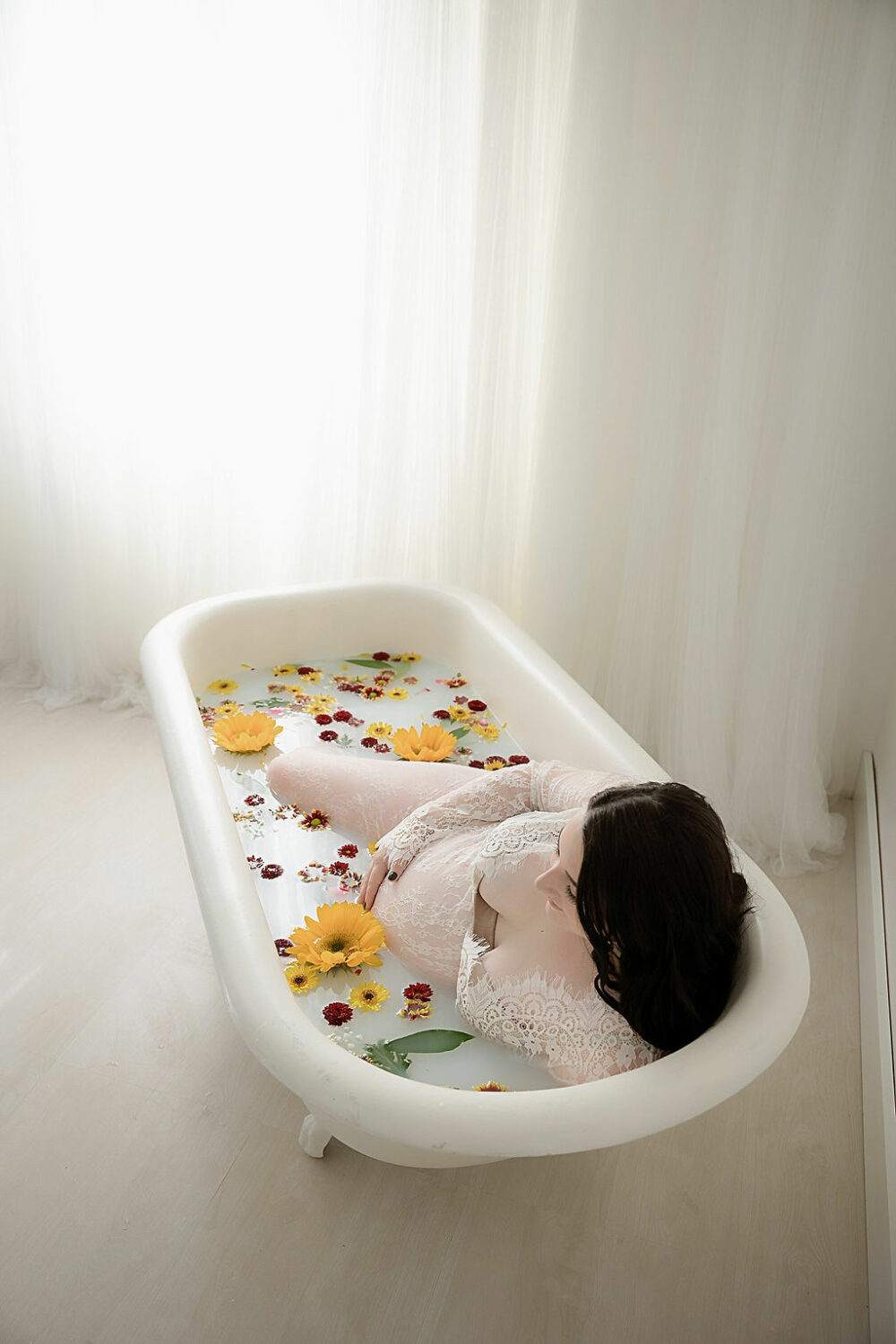 Woman laying in milk bath adorned with flowers wearing maternity gown for her maternity photo shoot poses taken in studio in Camden, New Jersey.