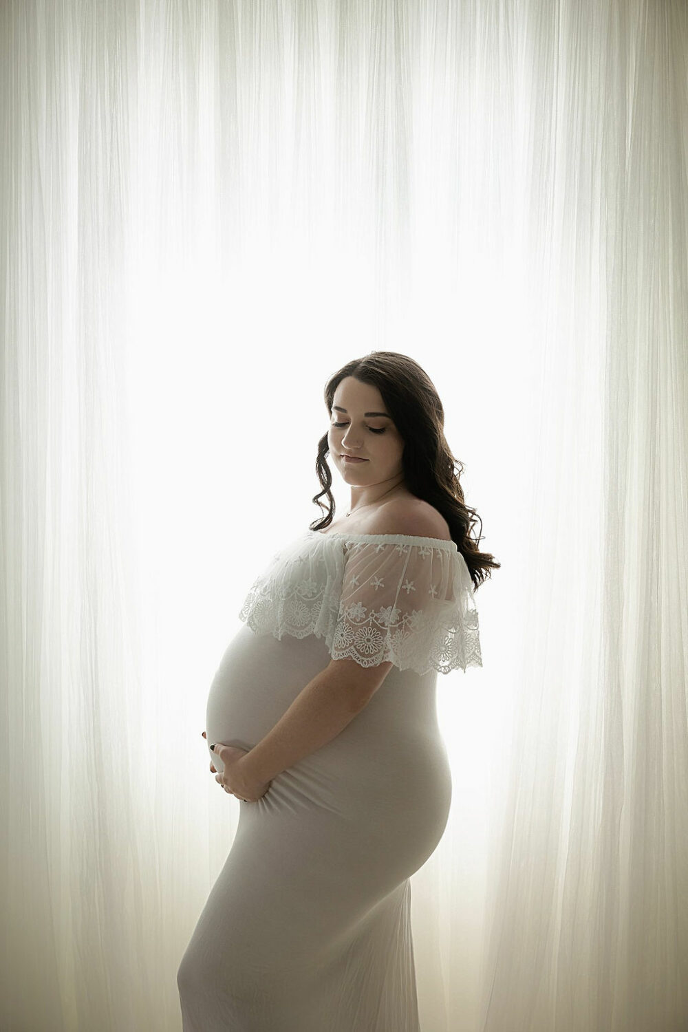 Woman standing and posing for her pregnancy pictures wearing white maternity gown again say light and bright background for her bra maternity and newborn session in Westhampton, New Jersey.