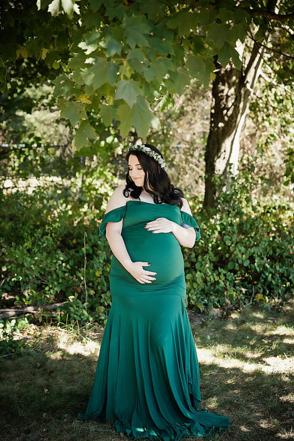 Woman standing outdoors in front of green foliage holding her belly and posing in a maternity gown for her maternity pictures taken in Southampton, New Jersey.