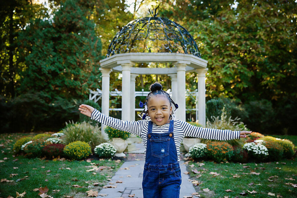 Young girl smiling and running towards camera standing in front of gazebo for her outdoor for an outdoor fall foliage family session in Sayen Gardens, New Jersey