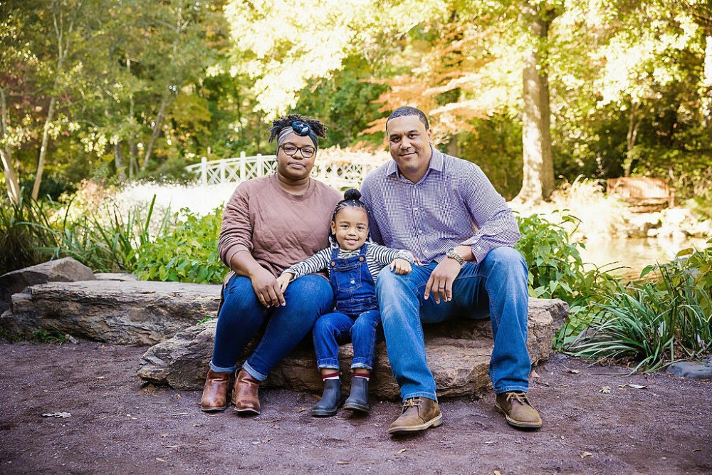 Man and woman with their young daughter sitting outdoors on a rock smiling for their fall foliage family session taken in freehold, New Jersey.