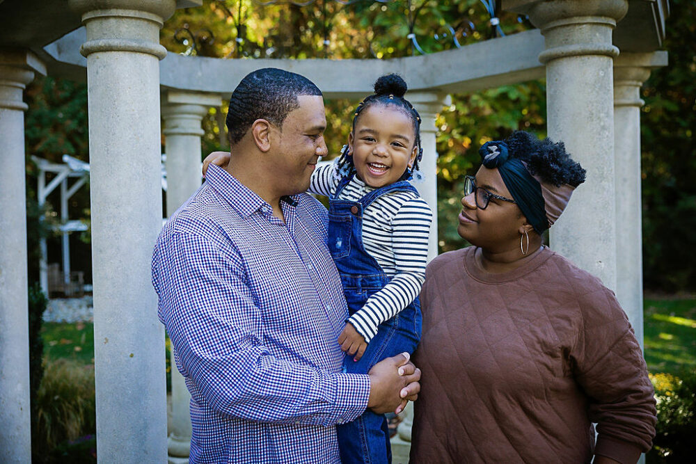 Man and woman looking at their daughter as she smiles at the camera and is in her fathers arms with a gazebo in the background for their fall foliage family session taken in Cherry Hill, New Jersey.