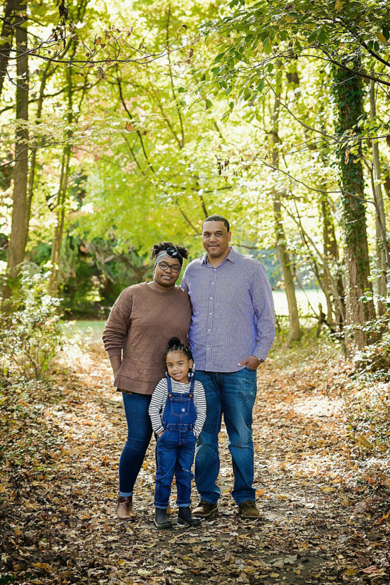 Man and woman standing next to each other and half embracing, and their young daughter standing in front of them all smiling for the camera for a Fall Foliage Family Session taken in Mount Holly, New Jersey.