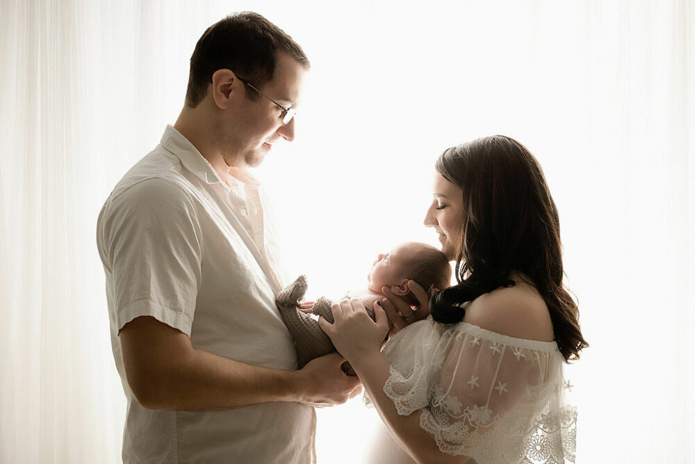 Man and woman looking at each other as they hold their newborn in their arms in between them and smiling standing against a light and bright background for their newborn photography session in Haddonfield, NJ.