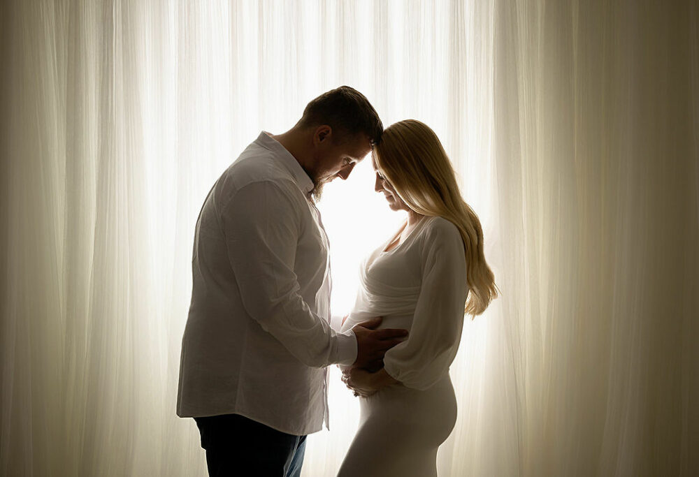 Man and woman touching foreheads as they both hold woman's belly for their couple maternity poses taken in professional studio in Manville, New Jersey.