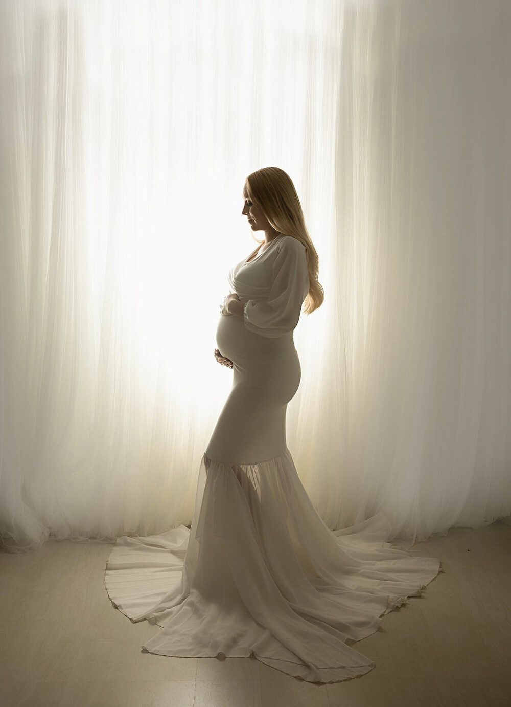 Woman standing and facing left while doing pregnant posing and holding her belly against a light background for her in studio maternity session take in Branchburg, New Jersey.