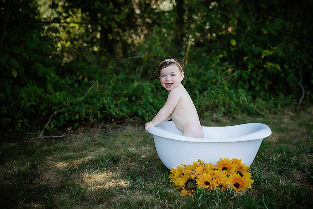 One-year-old girl smiling a camera she stands in bathtub photography prop adorned with flowers and matching headband for sunflower first birthday session in Westampton, New Jersey.