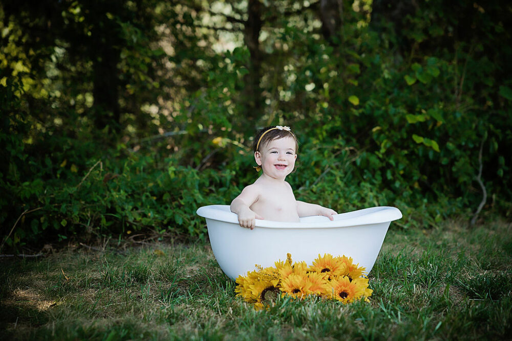 Young girl sitting in milk bath adorned with flowers, smiling at camera for her baby photos taken outside for her sunflower first birthday session in Camden, New Jersey.