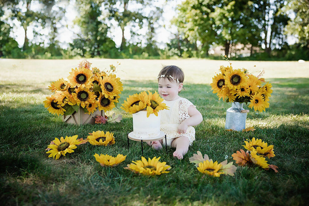 Toddler girl sitting on grass outdoors looking at her cake for her sunflower first birthday session in Hamilton, New Jersey.