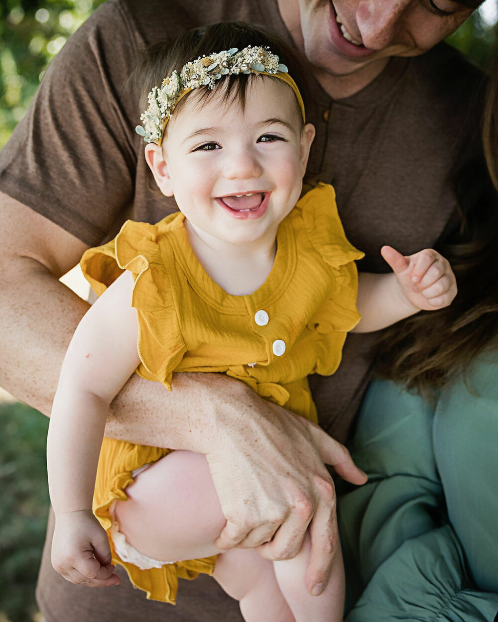 Close-up of toddler girl smiling in her fathers arms wearing overall outfit and headband for her sunflower first birthday session Pemberton, New Jersey.