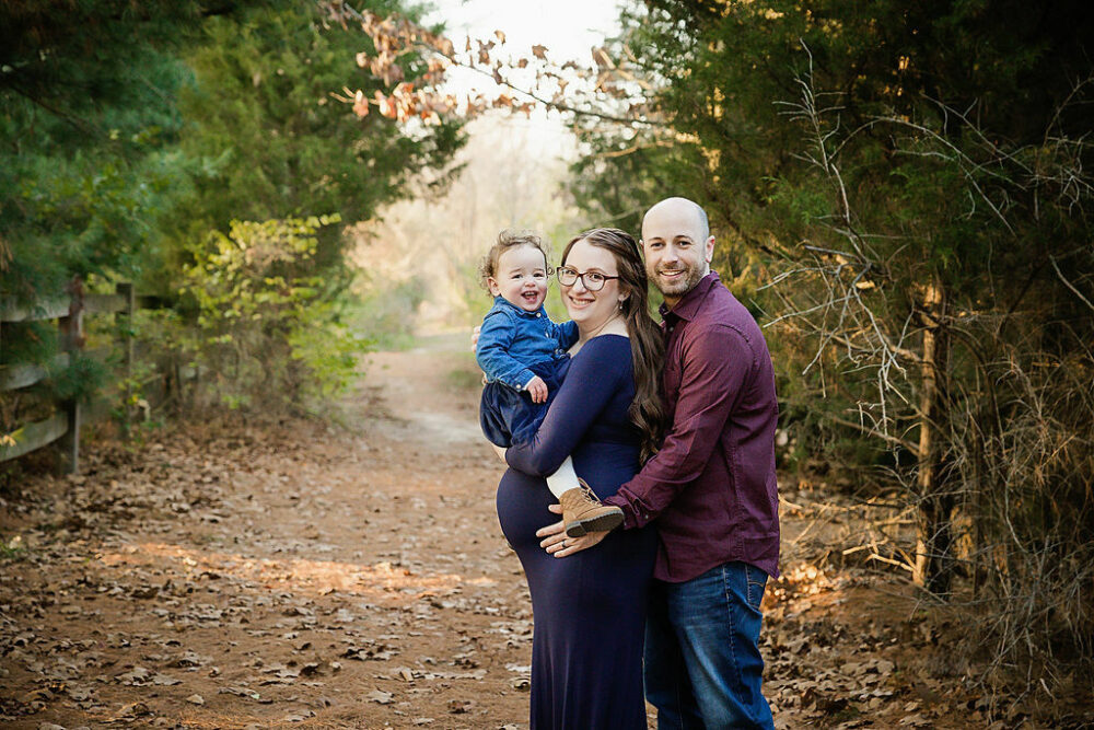 Family portrait of a woman and man and their toddler, all looking at the camera while he stands behind his wife and holding her belly for their navy maternity session taken in Burlington, New Jersey.
