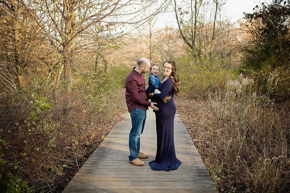 Man and woman standing on wooden trail in the park, standing across from each other, while holding their toddler boy for her Navy Maternity Session taken at Boundary Creek, New Jersey.