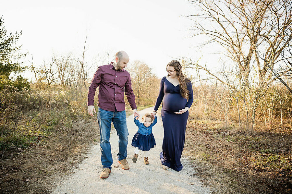 Lifestyle, photo of a man and a woman holding toddler girls hand as a walk on Park Trail for their outdoor navy maternity session, taken in Eastampton, New Jersey.